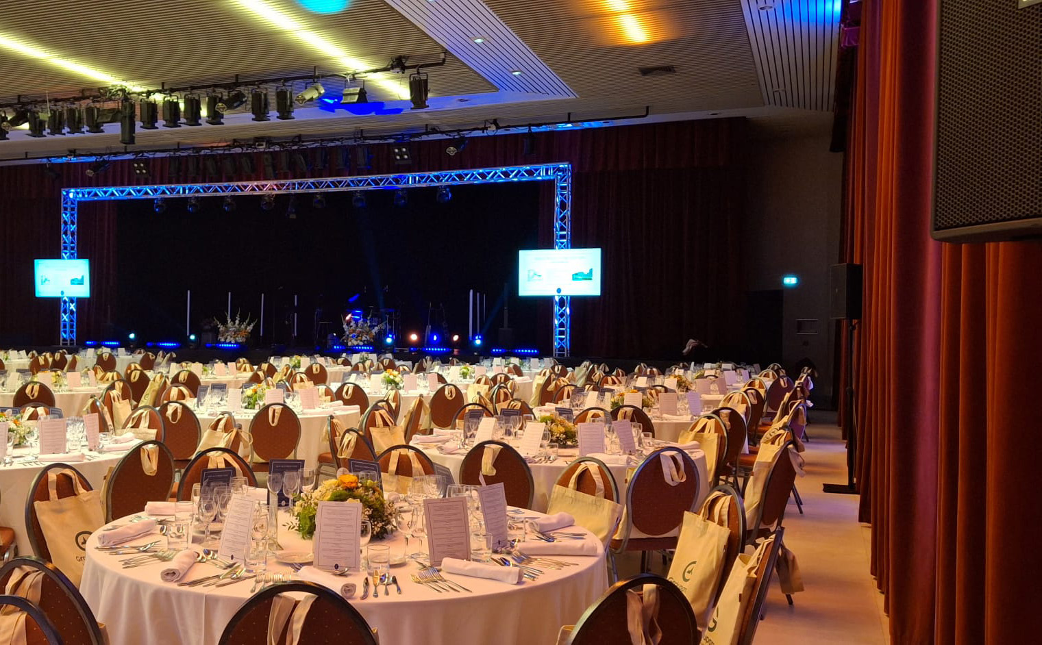 sonorisation_site Salle de réception avec tables rondes dressées, scène équipée en sonorisation et éclairage professionnel pour événement d’entreprise ou gala