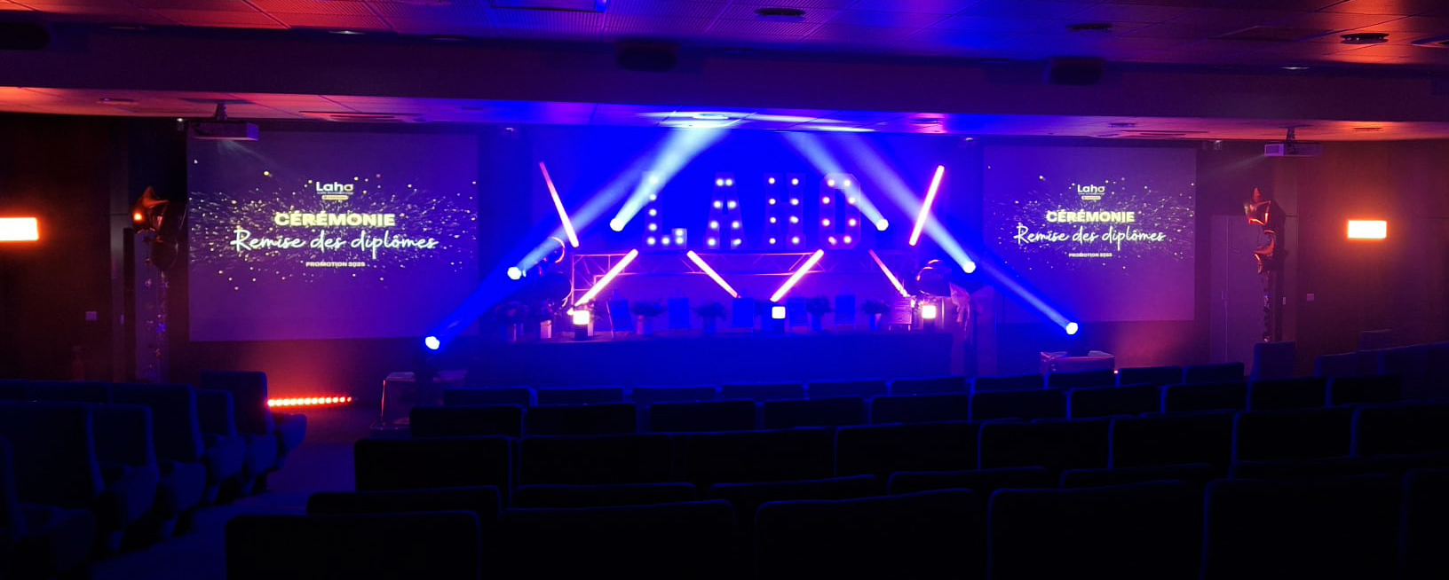 vidéo_site Scène de la cérémonie de remise des diplômes Laho Formation avec jeux de lumières bleus et violets, lettres géantes lumineuses et projection vidéo sur 2 toiles entourant la scène.